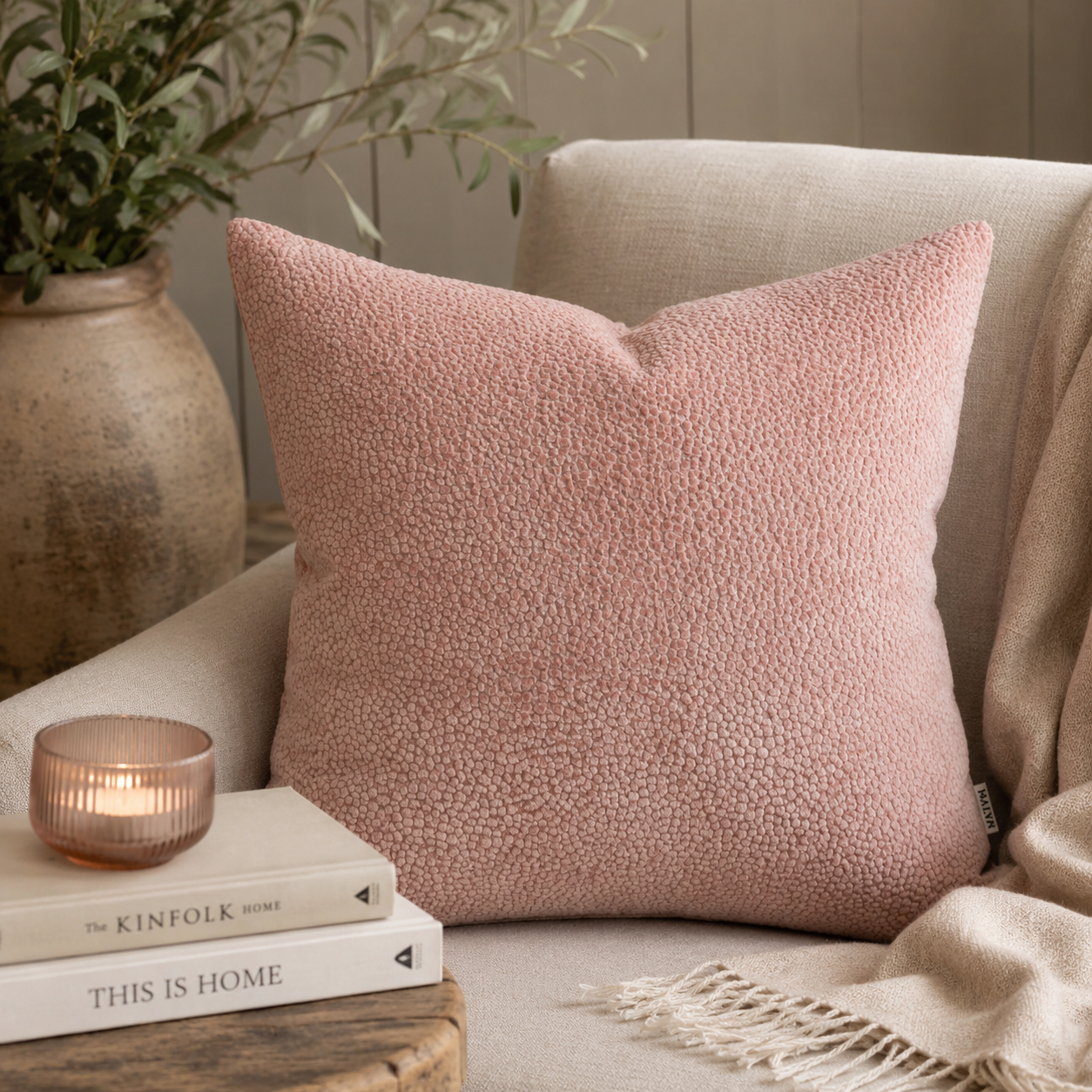 A dusty pink textured throw pillow displayed on a beige armchair in a cozy living room setting, styled with a lit candle, books, and a throw blanket.
